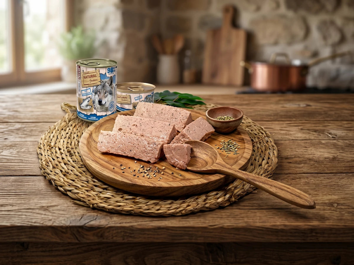 Una detallada fotografía de producto de alta calidad ambientada en una cocina de estilo rústico. En el centro, una tabla de madera redonda sobre un mantel de mimbre trenzado contiene varios bloques de paté de carne de comida para mascotas cortados. A la izquierda, se muestran dos latas de comida húmeda de la marca "Natural Greatness": una grande con la ilustración de la cara de un lobo y otra pequeña con ilustraciones de salmones. Una cuchara de madera grande, hojas verdes frescas y granos de pimienta negra