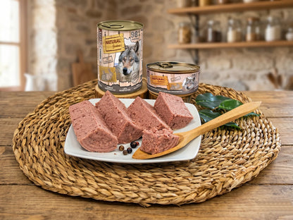 Fotografía realista de presentación de producto Natural Greatness Conejo. Se muestran las dos latas (400g y 200g) junto a un plato blanco con el paté de conejo cortado en rodajas, mostrando su textura carnosa. Todo está dispuesto sobre un mantel de mimbre en una cocina de estilo rústico y luminoso.